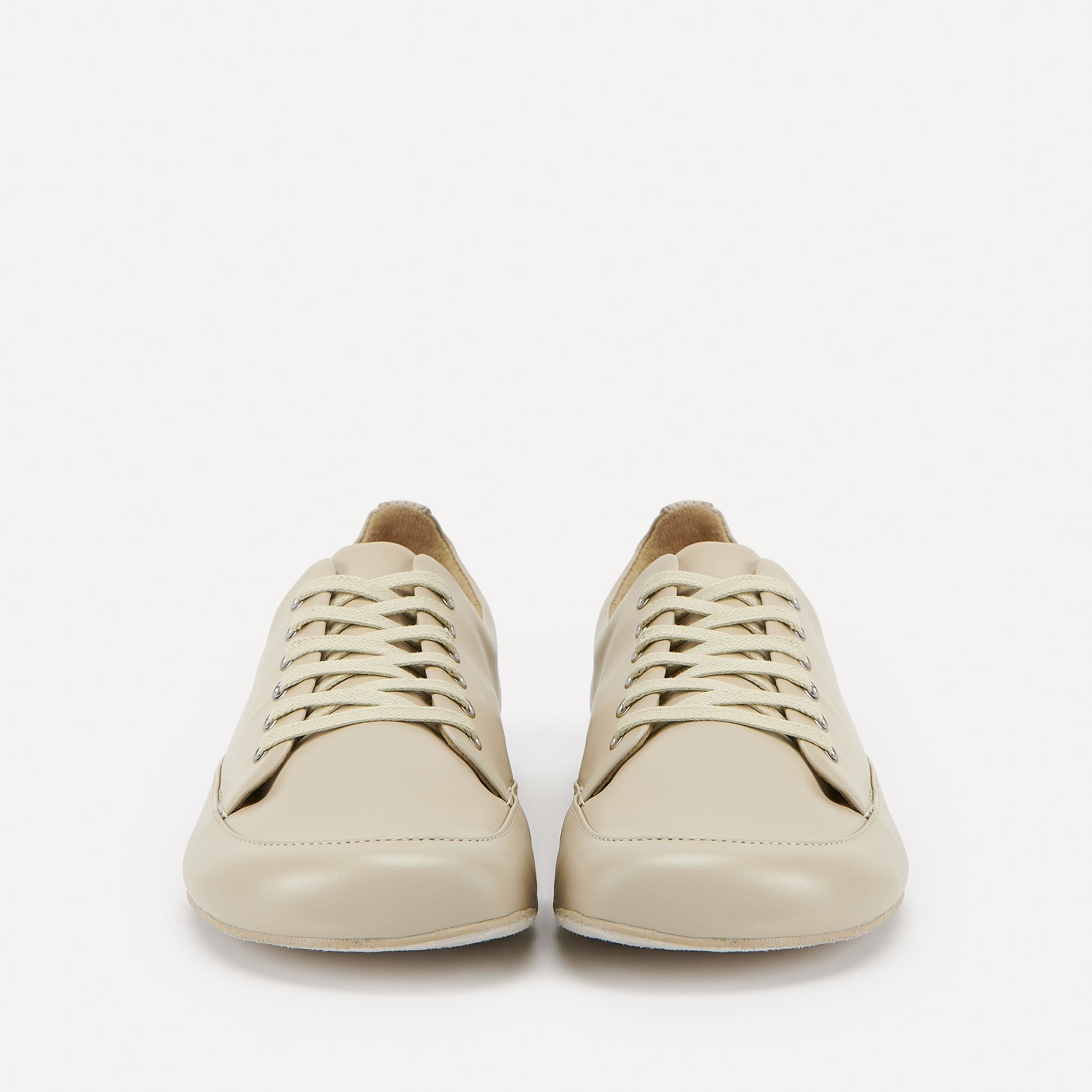 A pair of cream-colored leather lace-up barefoot dance sneakers with beige laces and silver eyelets, displayed on a clean off-white background.