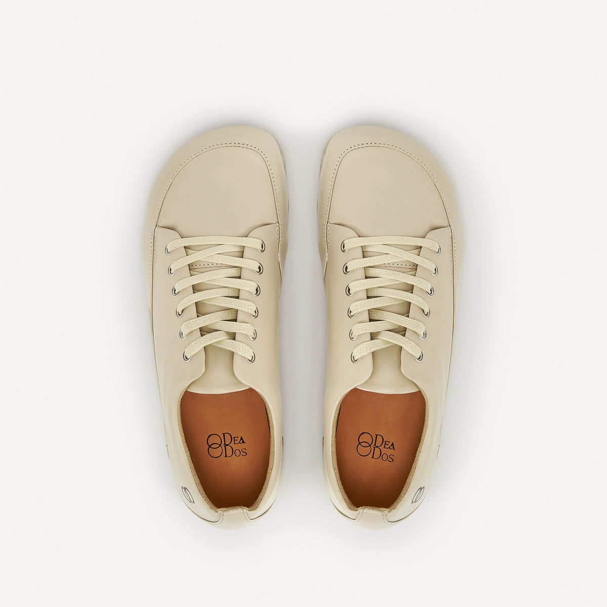 A pair of cream-colored leather lace-up barefoot dance sneakers with beige laces, silver eyelets and a black logo saying "DeaDos" on tan colored lining, shown from a top-down view on a clean off-white background.