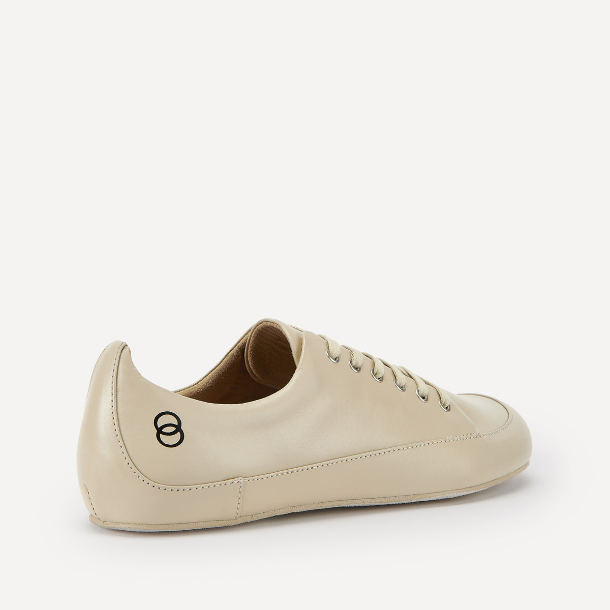 A single beige leather low-top barefoot dance sneaker shown from a side-rear angle on a clean, light off-white background. The shoe features a black double-ring logo on the heel, matching beige laces, and a  suede leather sole with a cream-colored painted rim.