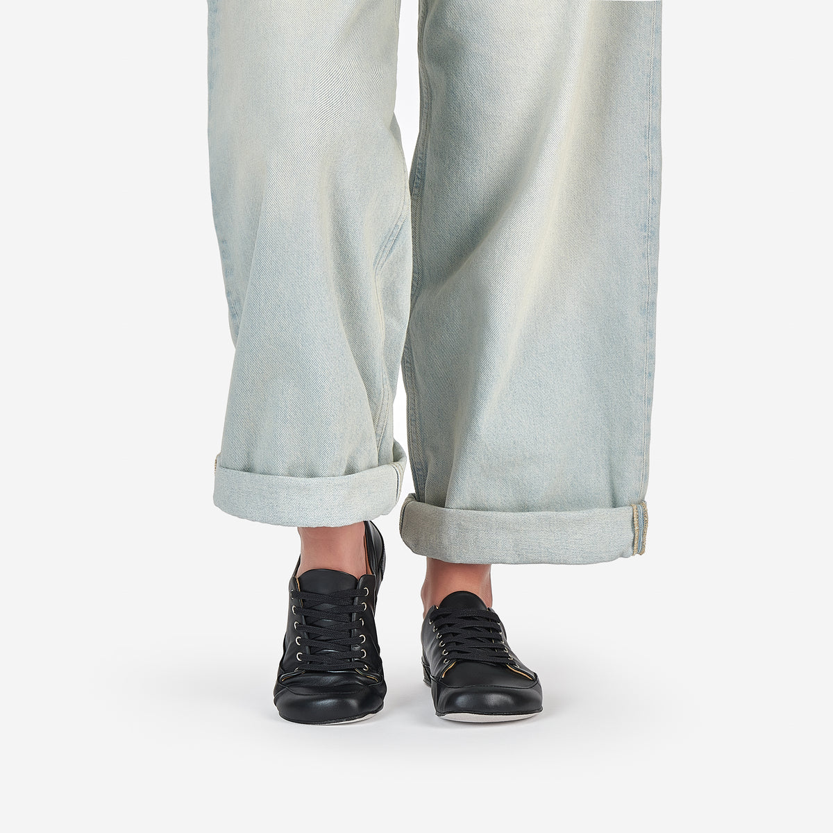 A person wearing black low-top barefoot dance sneakers and light-wash wide-leg jeans with rolled cuffs, shown from the knees down on a clean white studio background.