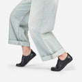 Black leather barefoot dance shoes worn by a  dancer in releve position, wearing light-wash wide-leg jeans, shown from the calves down against a clean white studio background.