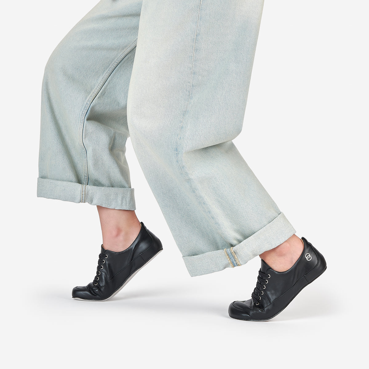 Black leather barefoot dance shoes worn by a  dancer in releve position, wearing light-wash wide-leg jeans, shown from the calves down against a clean white studio background.