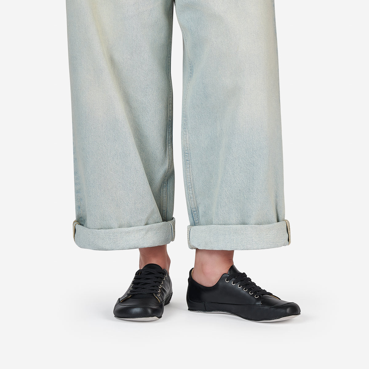 A person wearing black low-top barefoot dance sneakers and light-wash wide-leg jeans with rolled cuffs, shown from the knees down on a clean white studio background.
