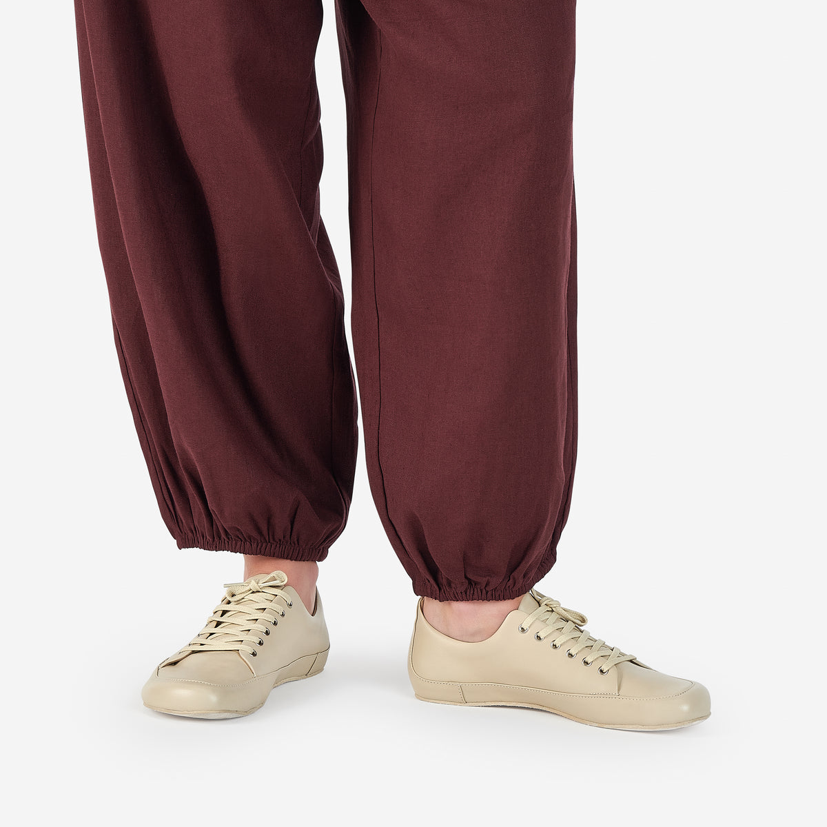 Close-up of a person wearing light beige low-top barefoot dance shoes in color cream, and loose dark burgundy harem-style pants, against a clean white studio background.