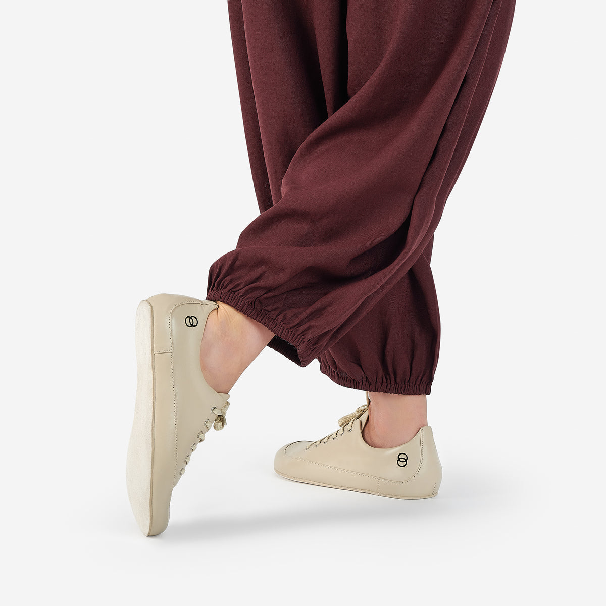 Close-up of a person wearing light beige low-top barefoot dance shoes in color cream, and loose dark burgundy harem-style pants, shown from the tights down, against a clean white studio background.