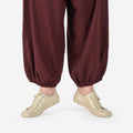 Close-up of a person on releve wearing light beige low-top barefoot dance shoes in color cream, and loose dark burgundy harem-style pants, against a clean white studio background.
