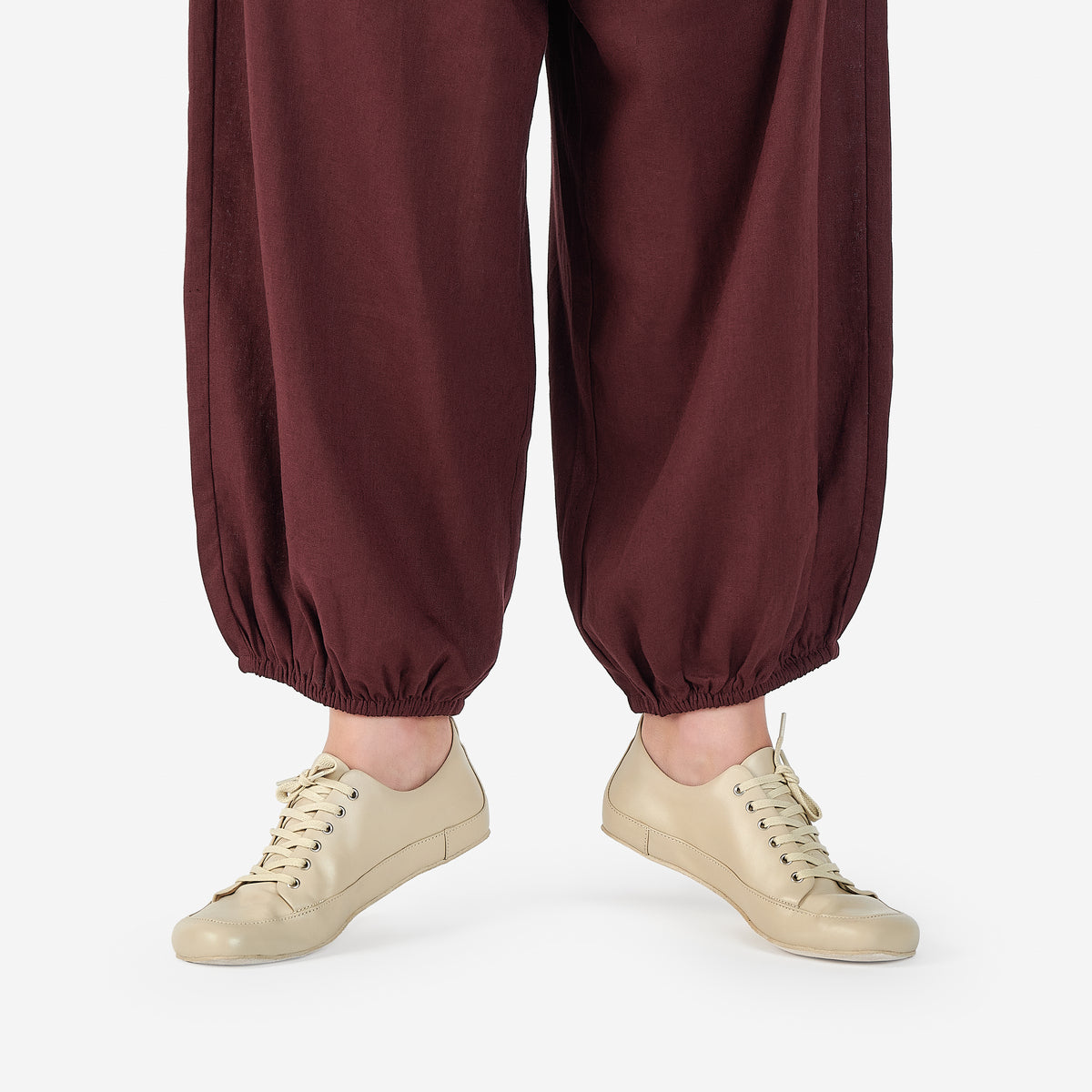 Close-up of a person on releve wearing light beige low-top barefoot dance shoes in color cream, and loose dark burgundy harem-style pants, against a clean white studio background.