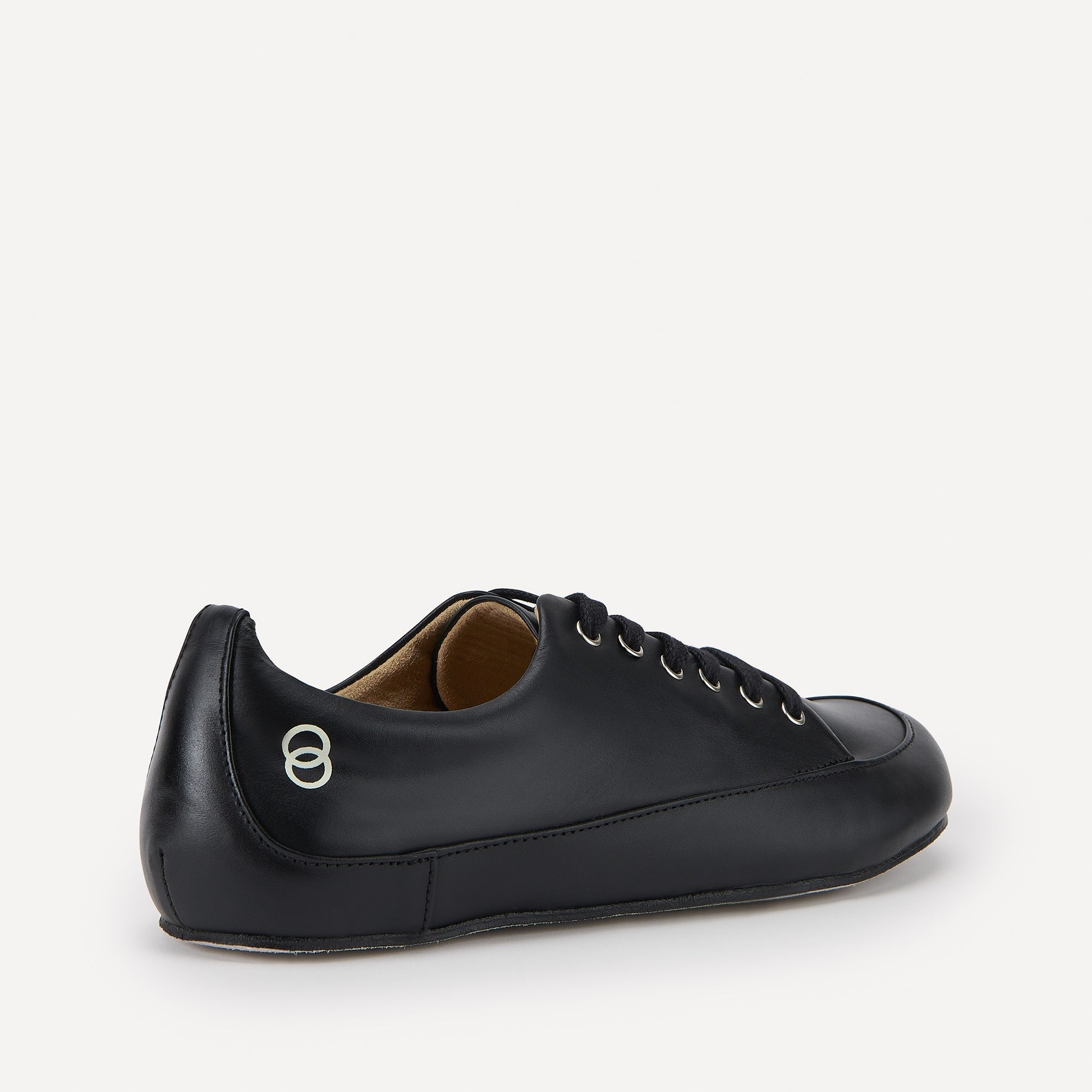 A black leather lace-up barefoot dance sneaker a beige colored logo showing two vertically overlapping rings, displayed on a clean off-white background.